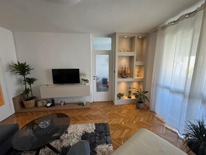 Una sala de estar con televisor de pantalla plana, un sofá, una mesa de centro y piso de madera con patrón de espiga. Hay plantas en macetas, una estantería con adornos y una puerta con vidrio esmerilado.