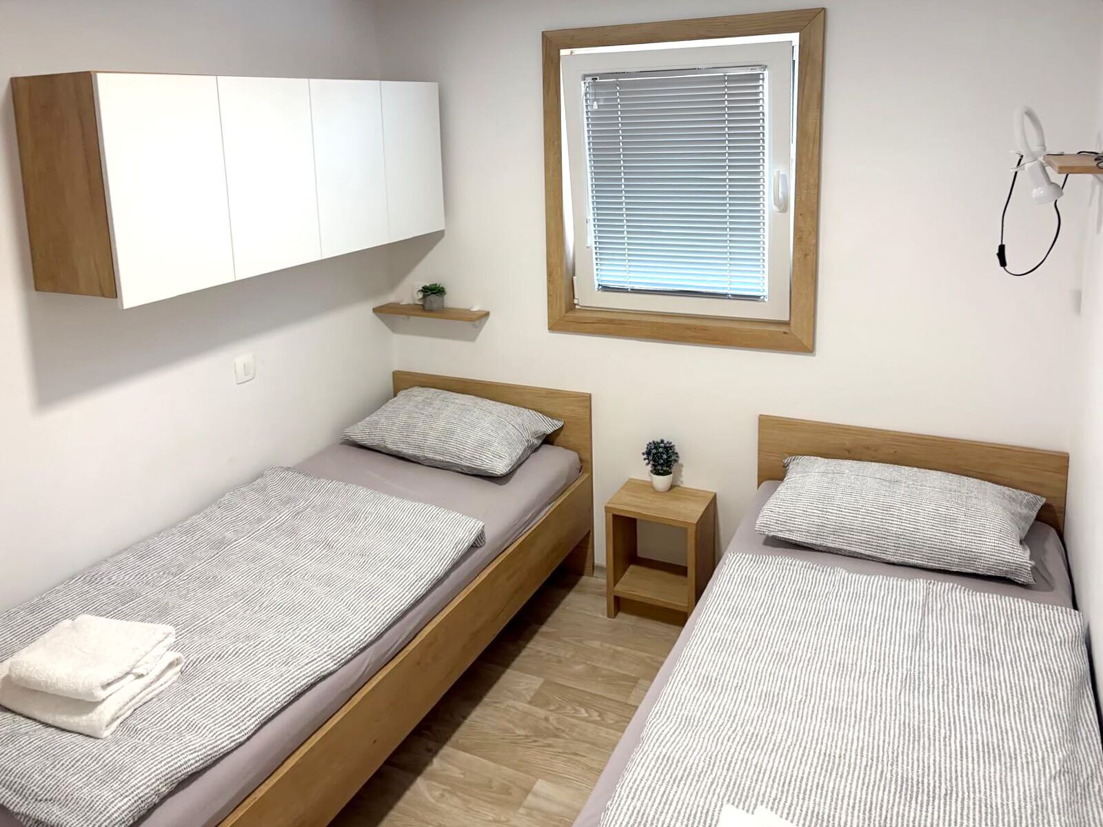 A bedroom with two single beds, a wooden wardrobe, a small bedside table with a plant, a window with blinds, and a wall-mounted lamp.