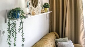 A cozy living room with a beige sofa, a small side table, a decorative mirror, wall-mounted sconces, and a potted plant. - House Neda (Marina)