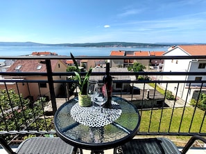 Ein Balkon mit rundem Tisch, einem Blumentopf und einer Flasche. Im Hintergrund ist ein Gewässer und Gebäude zu sehen.