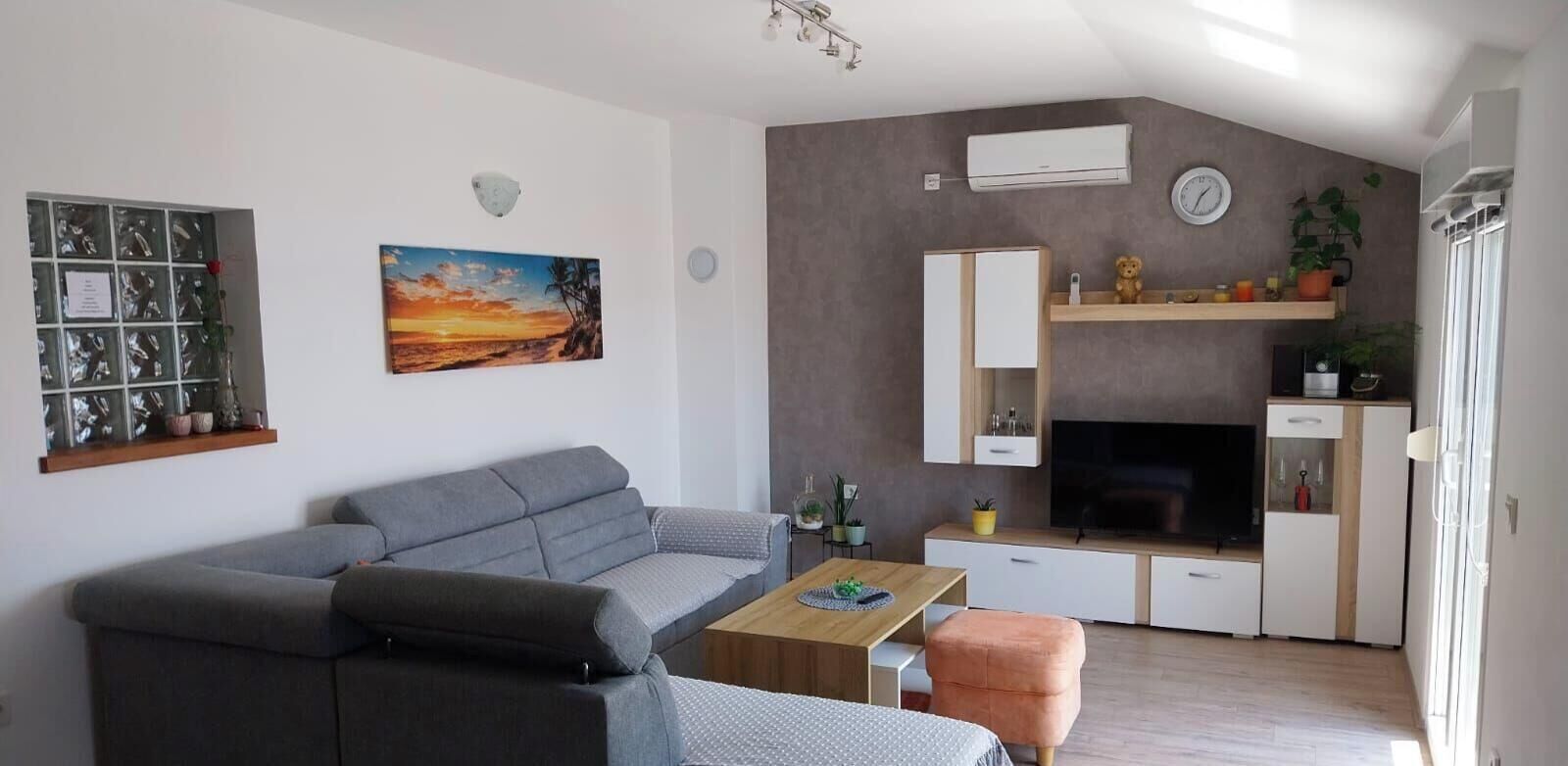 A modern living room with a grey sofa, a wooden coffee table, a small orange ottoman, a TV, and a fireplace.