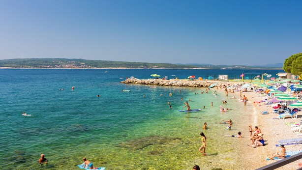 Una playa con agua cristalina, gente nadando y tomando el sol, y sombrillas.