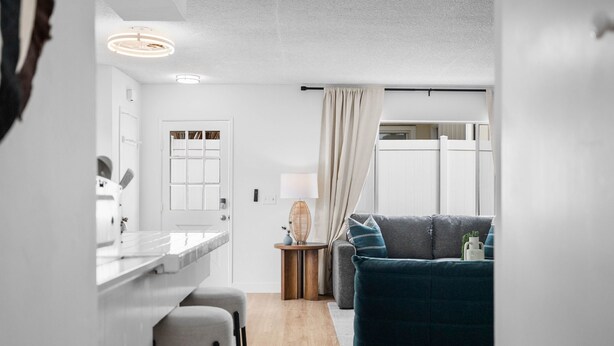 Eine moderne Küche mit weißer Arbeitsplatte und Barhocker, ein Essbereich mit dunkelblauem Sofa und einem kleinen Beistelltisch mit Lampe, sowie Blick auf eine weiße Tür und ein Fenster mit Vorhängen.