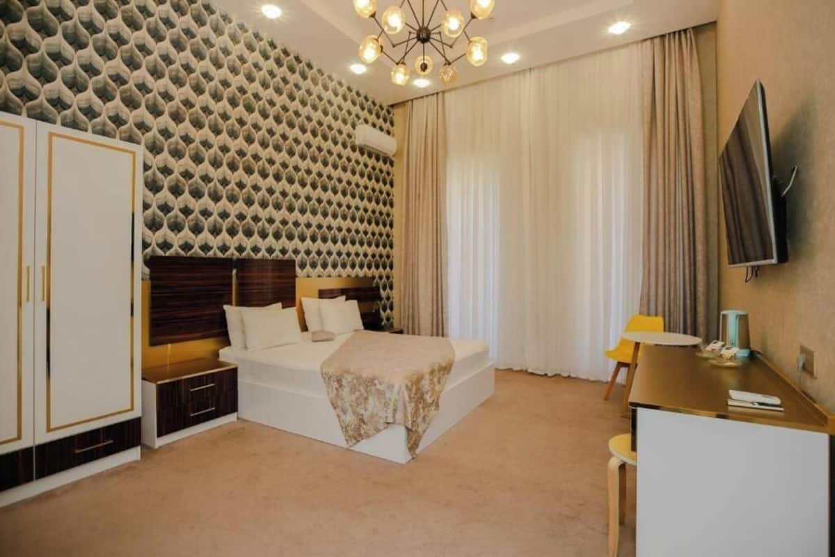 A hotel room with a patterned wall, a bed with a wooden headboard, a desk with a chair, and a flat-screen TV mounted on the wall.