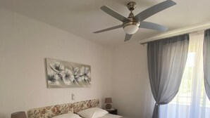 Een slaapkamer met een bed, twee nachtkastjes, een plafondventilator, een raam met gordijnen en een bloemschilderij aan de muur.