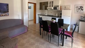 Een moderne woonkamer met een bank, eettafel en stoelen. In de achtergrond is een keuken zichtbaar met witte kasten en apparatuur.