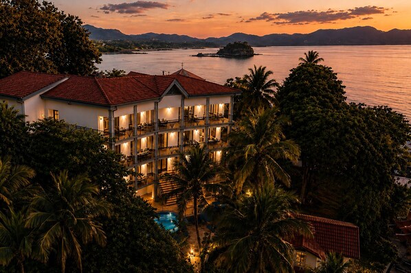 Ein mehrstöckiges Hotel mit Schwimmbad, umgeben von Palmen und mit Blick auf ein Gewässer mit einer entfernten Insel.