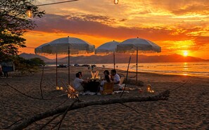 A beach scene with people dining under umbrellas, a sunset, and a distant mountain.