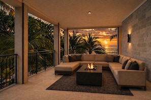 A modern living room with a large sectional sofa, a fireplace, and a view of the sunset over the ocean.