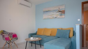 A living room with a blue sofa, a colorful chair, a small round table, and a painting of a lighthouse. - Apartments Alex (Rasa)