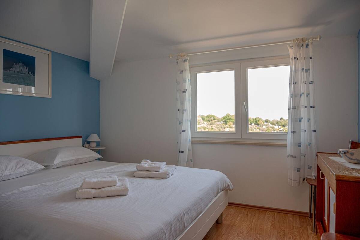 Une chambre à coucher avec un lit, une table de chevet, une lampe, une fenêtre avec des rideaux et une vue sur les arbres.