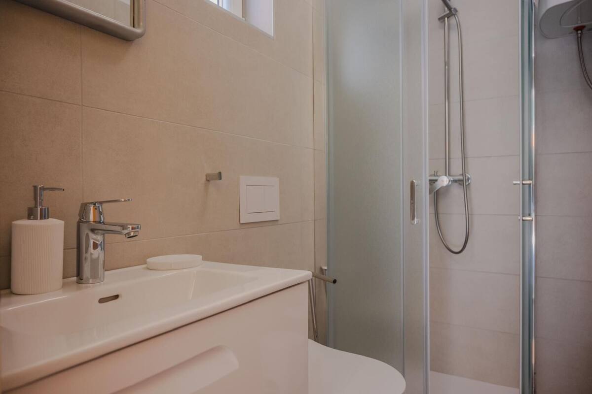 Une salle de bain moderne avec un lavabo blanc, un miroir, un distributeur de savon et une douche dotée d’une porte en verre.