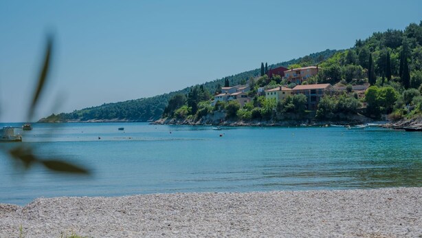A coastal area with a pebbled beach, a calm sea, and a hillside with buildings. - Apartments Alex (Rasa)