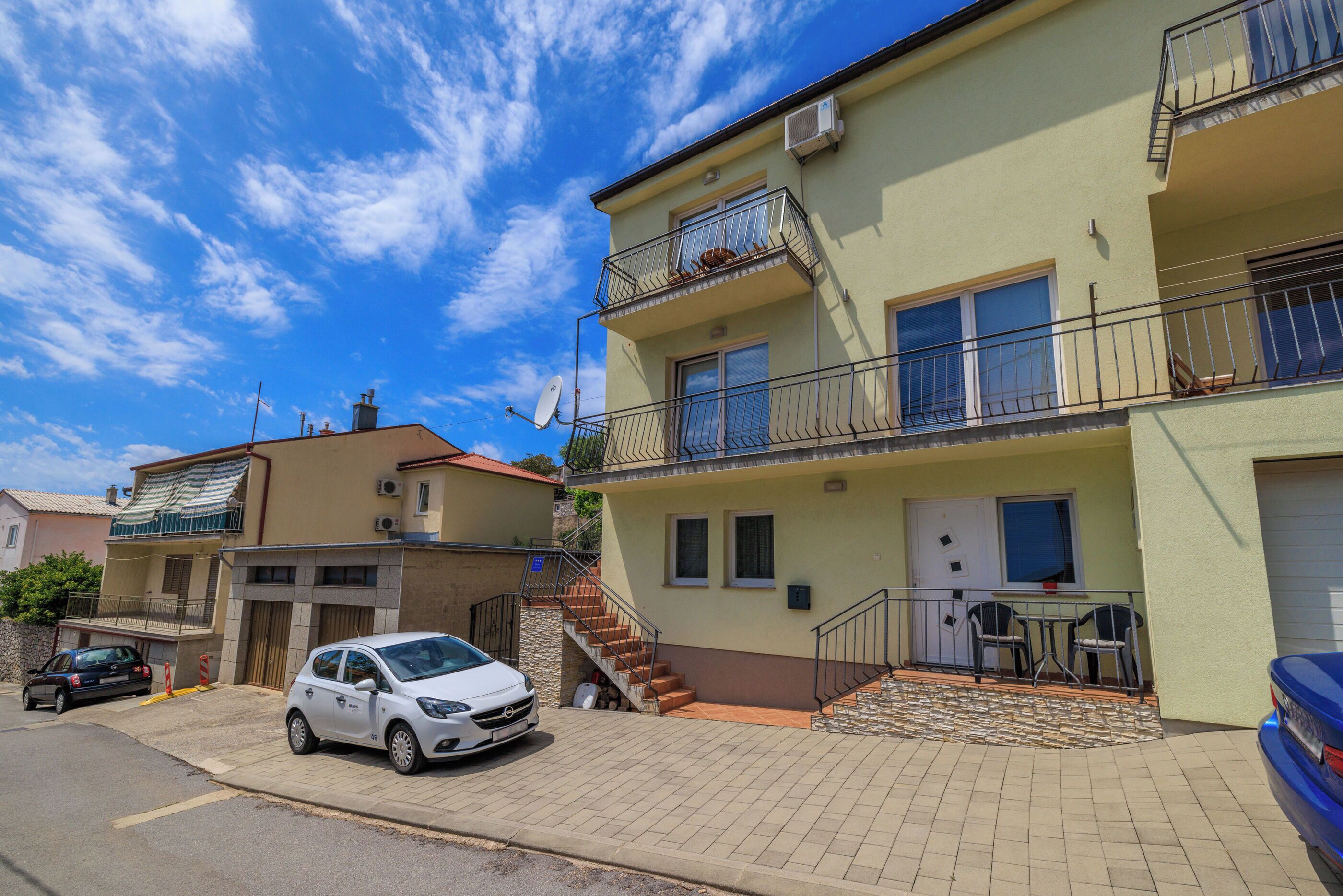 A residential building with a staircase leading to a balcony, a car parked in front, and a small outdoor seating area.