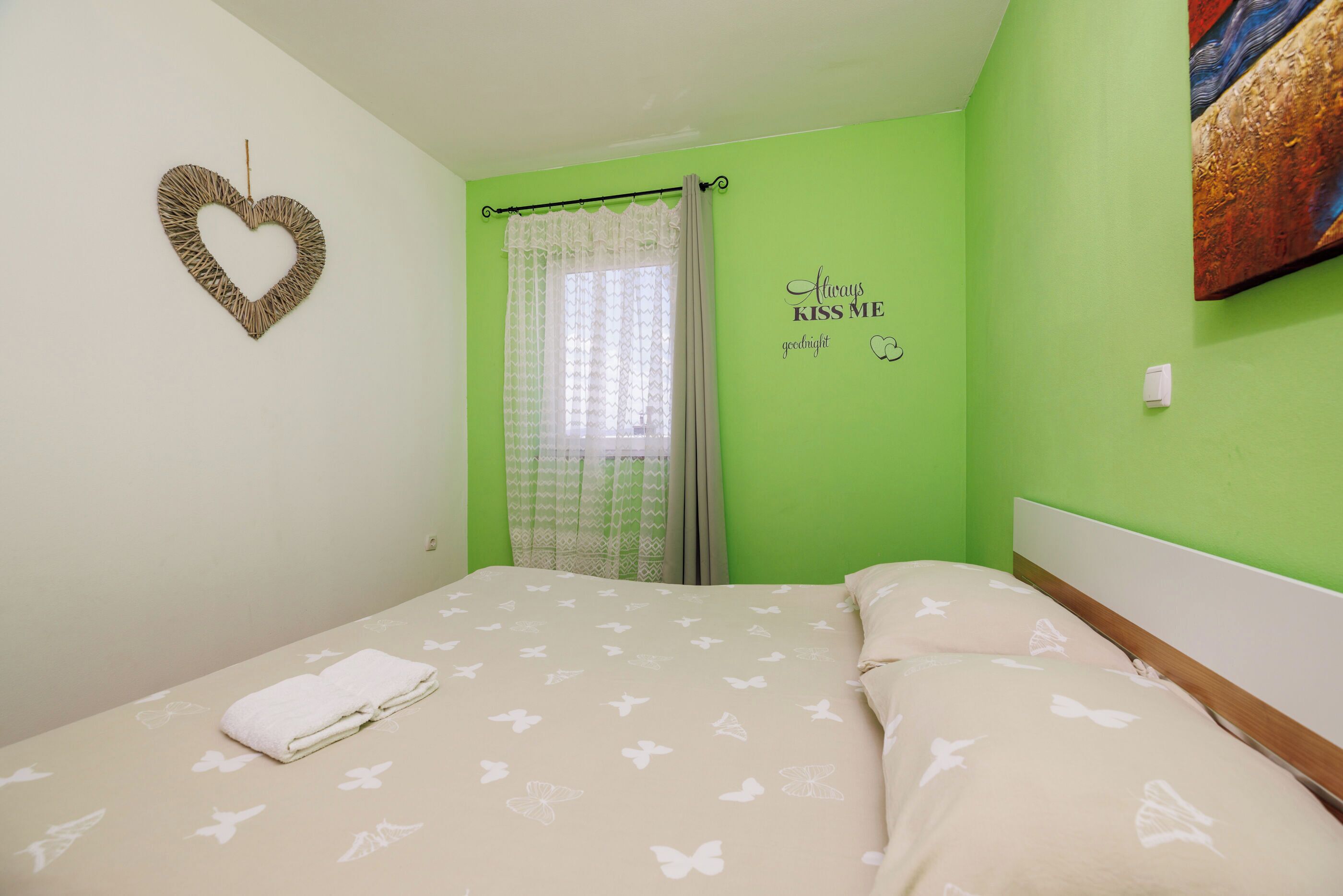 A bedroom with a bed, a heart-shaped decoration, a window with curtains, and a wall with a 'Always Kiss Me' sign.