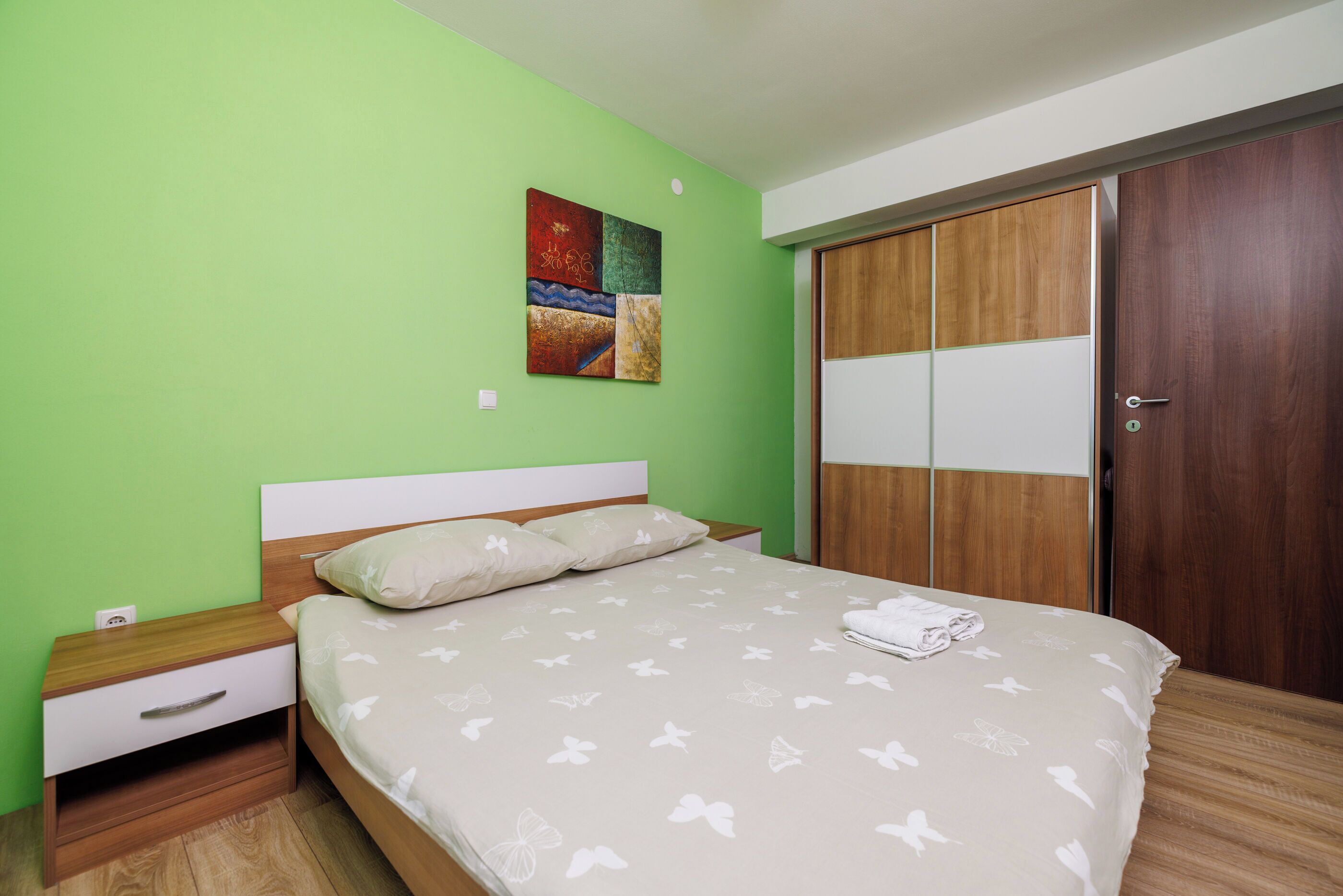 A bedroom with a bed, nightstands, a wooden wardrobe, and a colorful artwork on the wall.