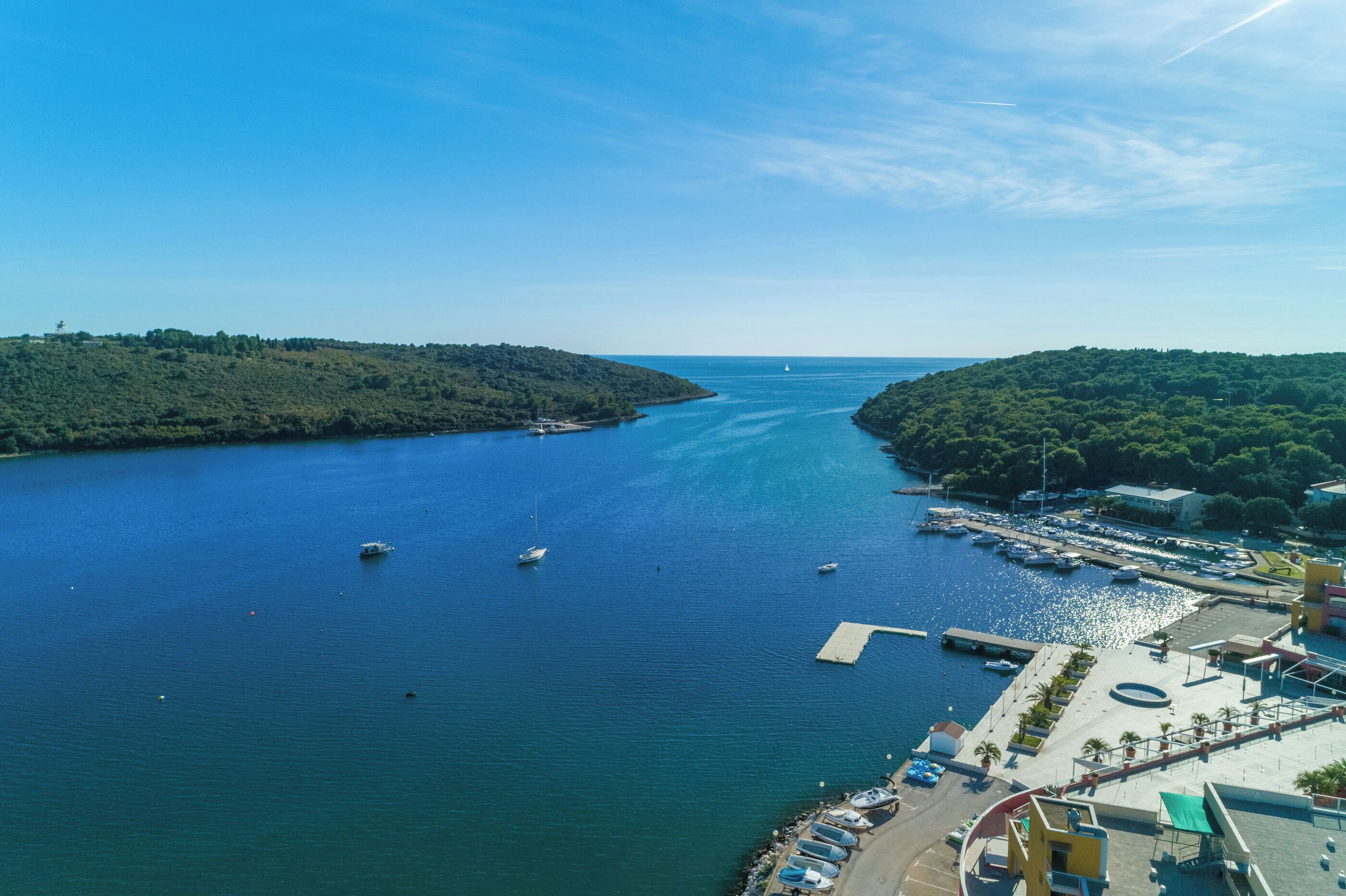 An aerial view of a marina with boats docked, a pier, and a hilly landscape in the background.