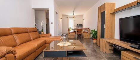 A modern living room with a leather sofa, a glass coffee table, a dining area with a wooden table and chairs, and a flat-screen TV mounted on a wooden cabinet.