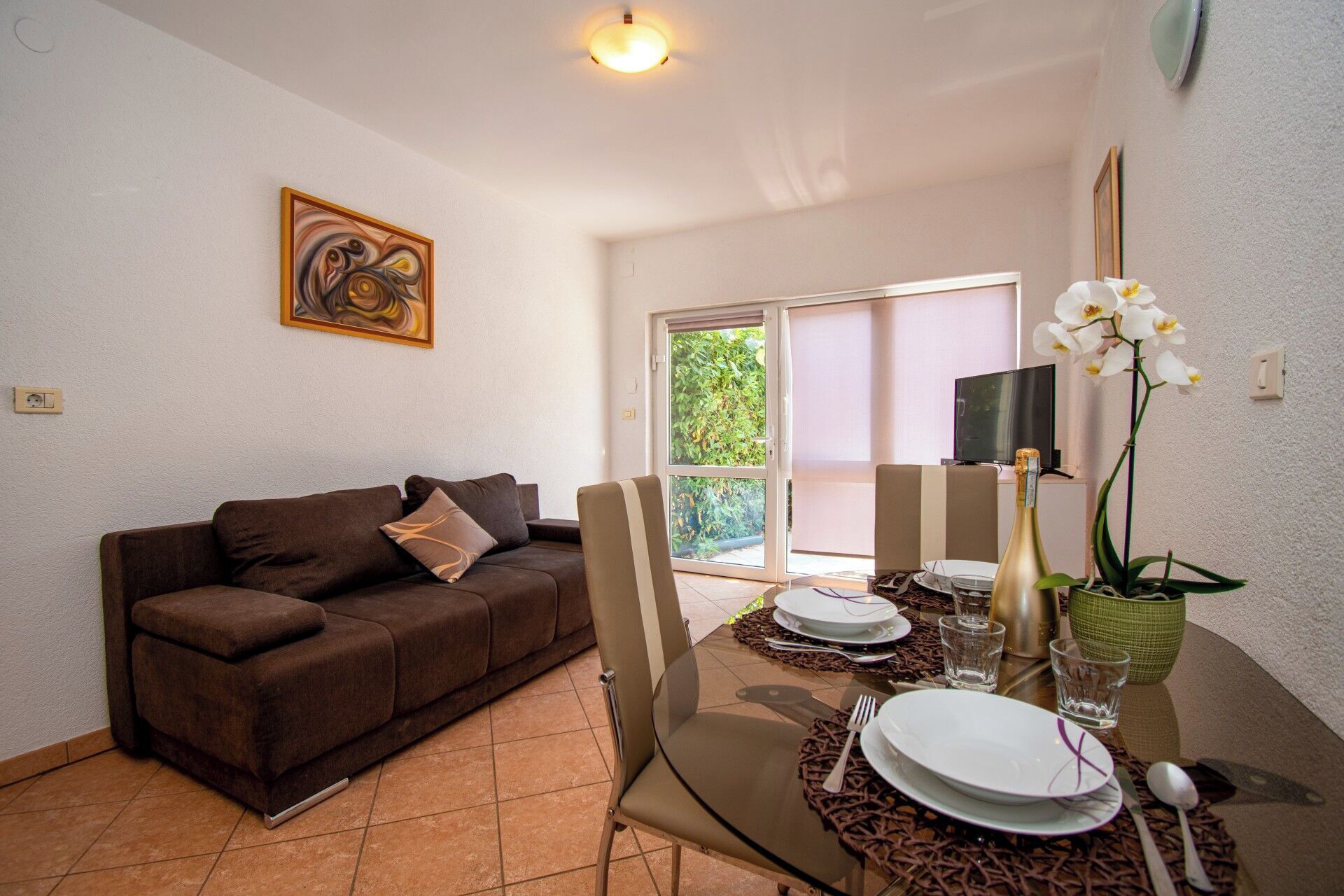 A living room with a brown sofa, a glass dining table set with plates, a bottle, and a vase with white flowers.