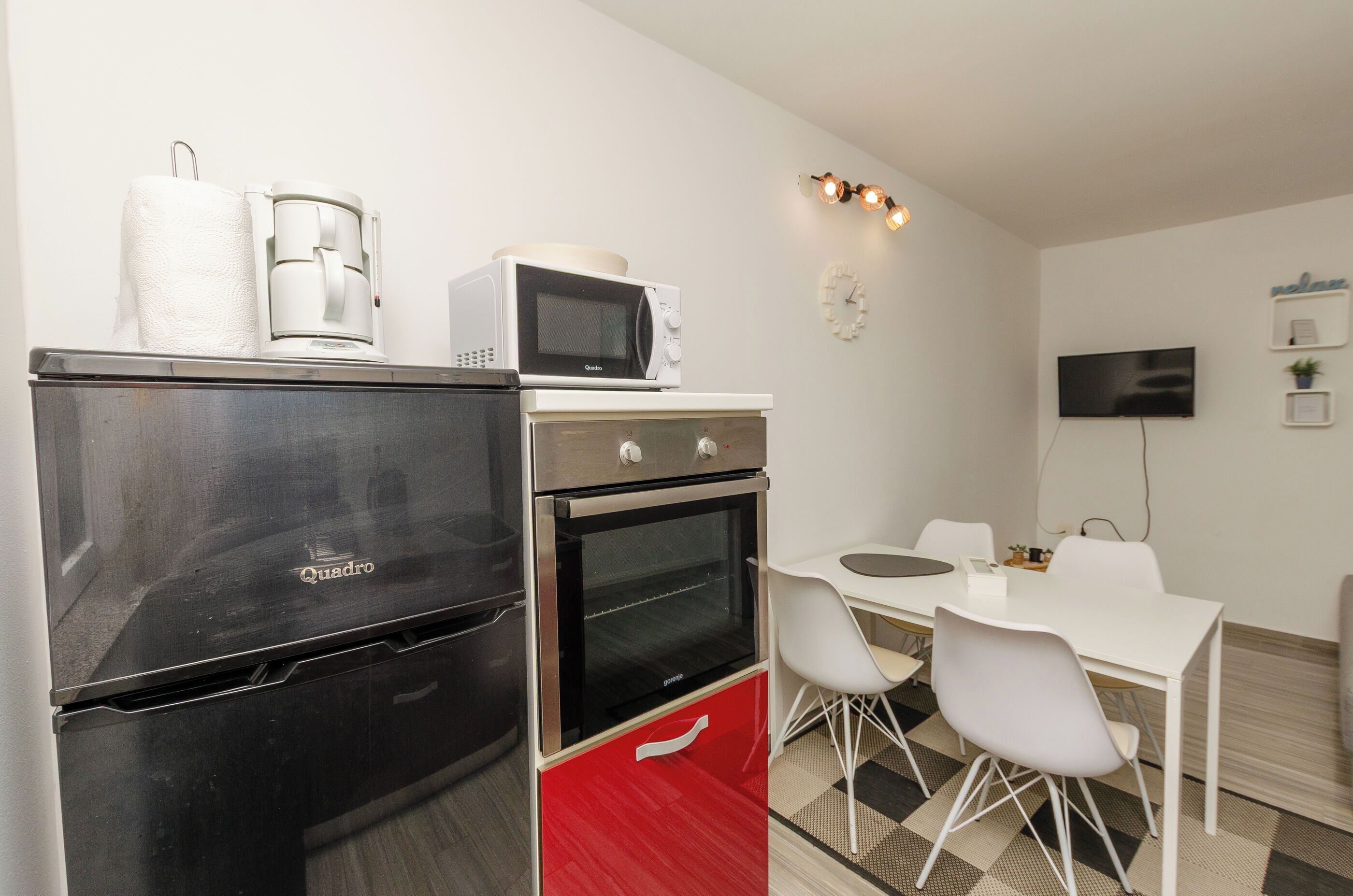 A compact kitchen with a refrigerator, microwave, and oven, a dining table with chairs, and a flat-screen TV mounted on the wall.