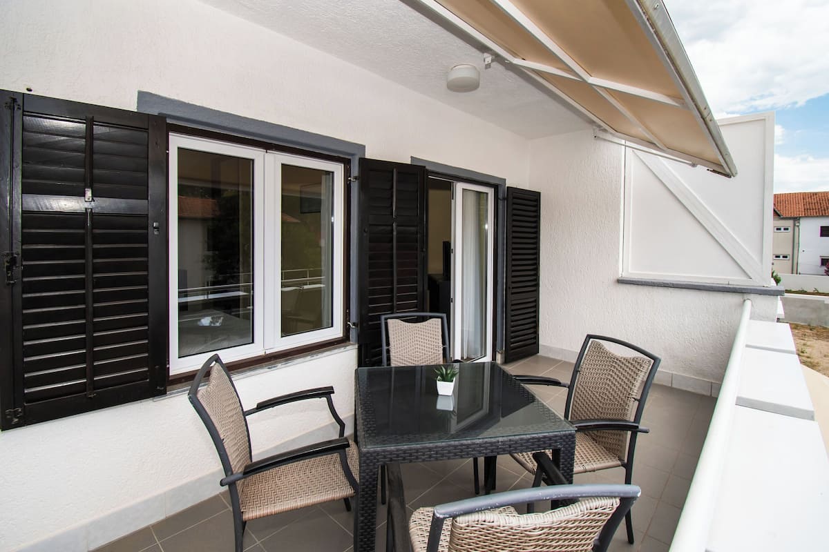 Une terrasse extérieure couverte, dotée d’une table en verre et de chaises en osier, et agrémentée de volets noirs aux fenêtres.