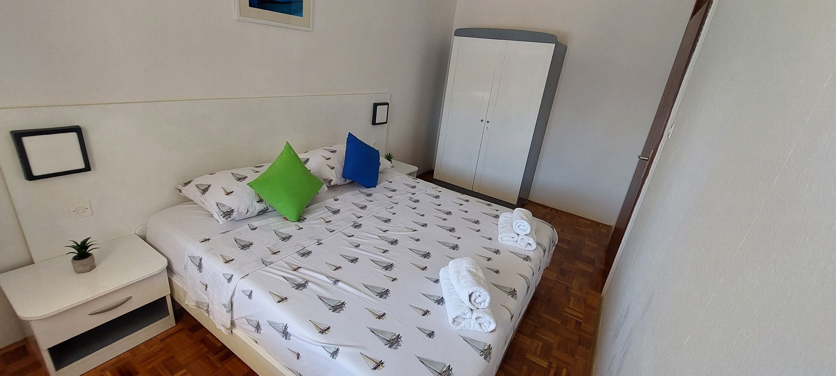 Un dormitorio pequeño y ordenado con cama, mesita de noche con planta, armario y un cuadro en la pared.