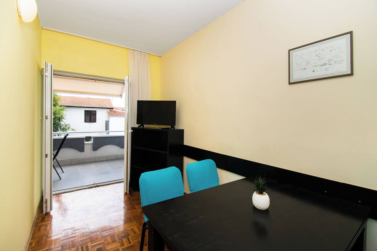 Une pièce avec une table noire, deux chaises bleues, une petite plante en pot, un tableau encadré au mur et un téléviseur posé sur un meuble.