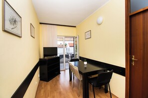 Une petite pièce étroite, avec un plancher en bois, une table noire et deux chaises, un téléviseur posé sur un meuble noir, et une porte donnant sur un balcon.