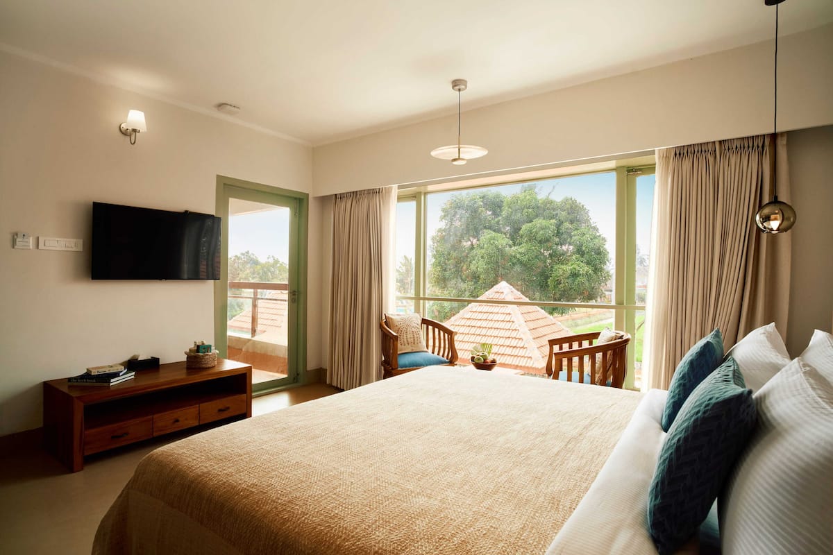 Un dormitorio amplio con una cama grande, un televisor en la pared, una silla de madera y un balcón con vistas a árboles.