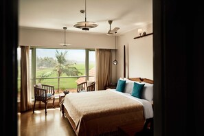 A hotel room with a large bed, a chair, a desk, and a view of a palm tree and greenery outside. - Vedic Village Retreat Alleppey (Alappuzha)