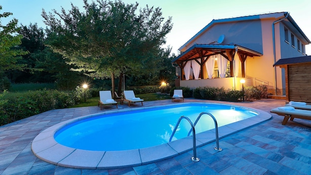 Ein Schwimmbad mit Liegestühlen, ein Haus mit überdachter Terrasse und ein Garten mit Bäumen und Sträuchern.