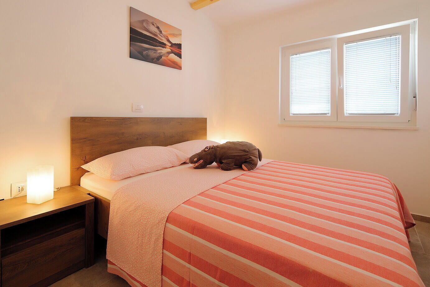 Ein Schlafzimmer mit einem hölzernen Kopfteil, einer gestreiften Bettdecke, einem Nachttisch mit Lampe und einem Fenster mit Jalousien.