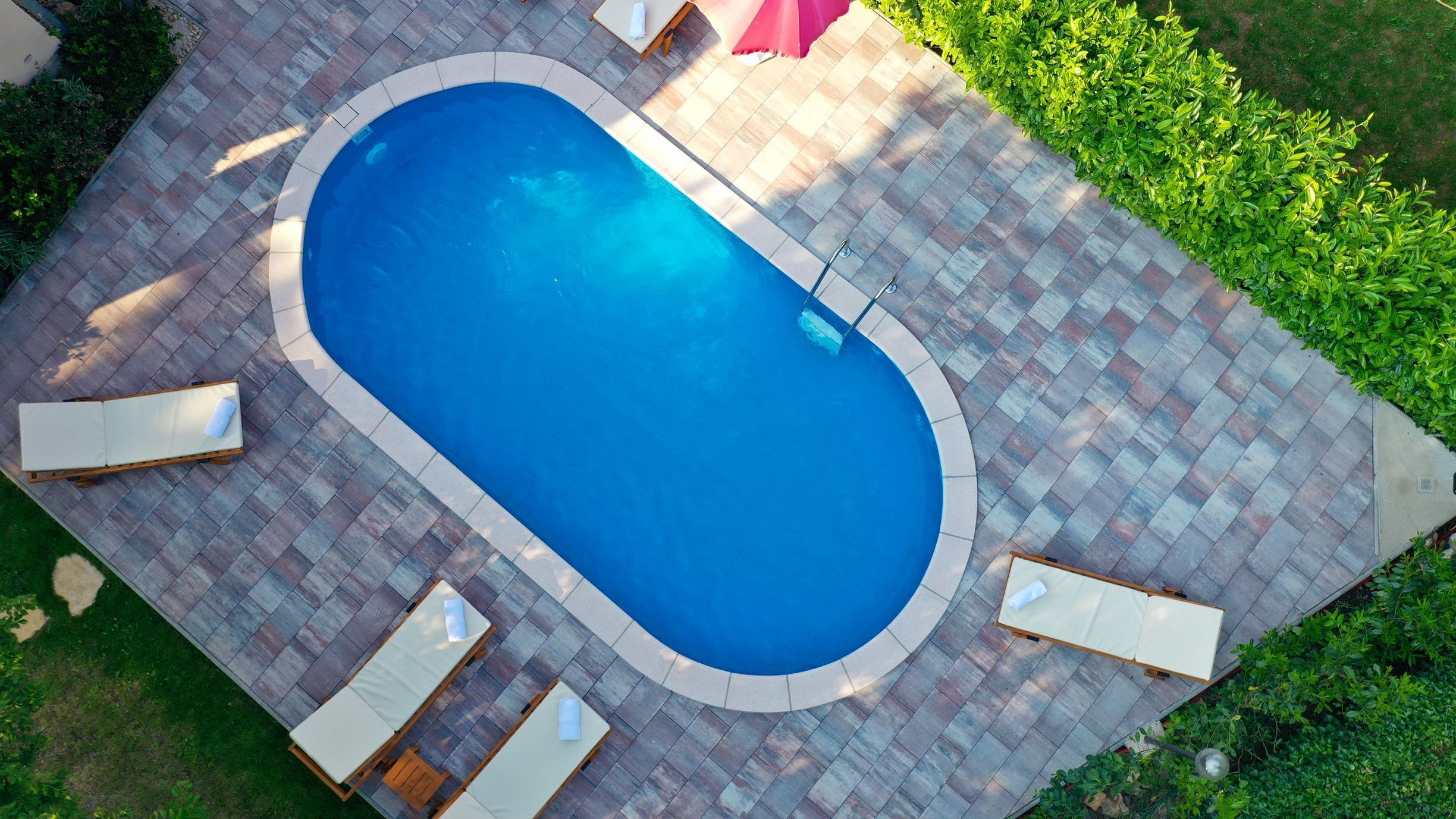 Et rektangulært pool med flisebelagt område, omgivet af liggestole og en lille parasol.