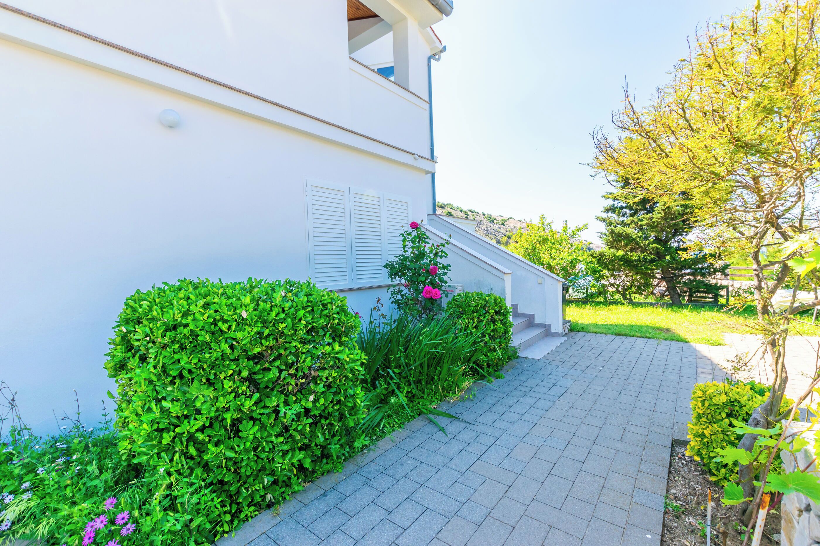 A pathway leading to a white building with a staircase and a small garden area with green bushes and pink flowers.