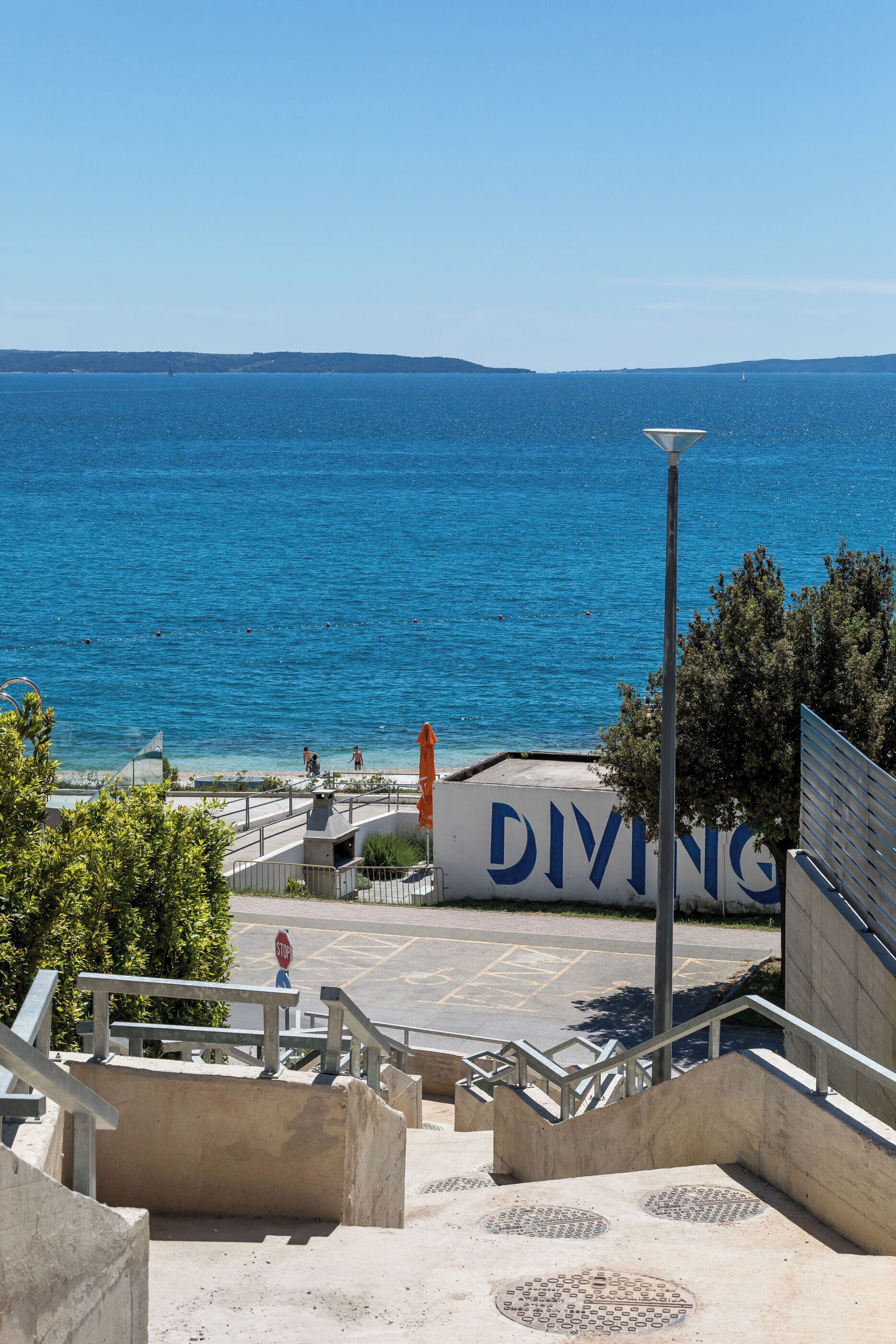 A coastal view with a diving sign, stairs leading to a beach, and a clear blue sea.