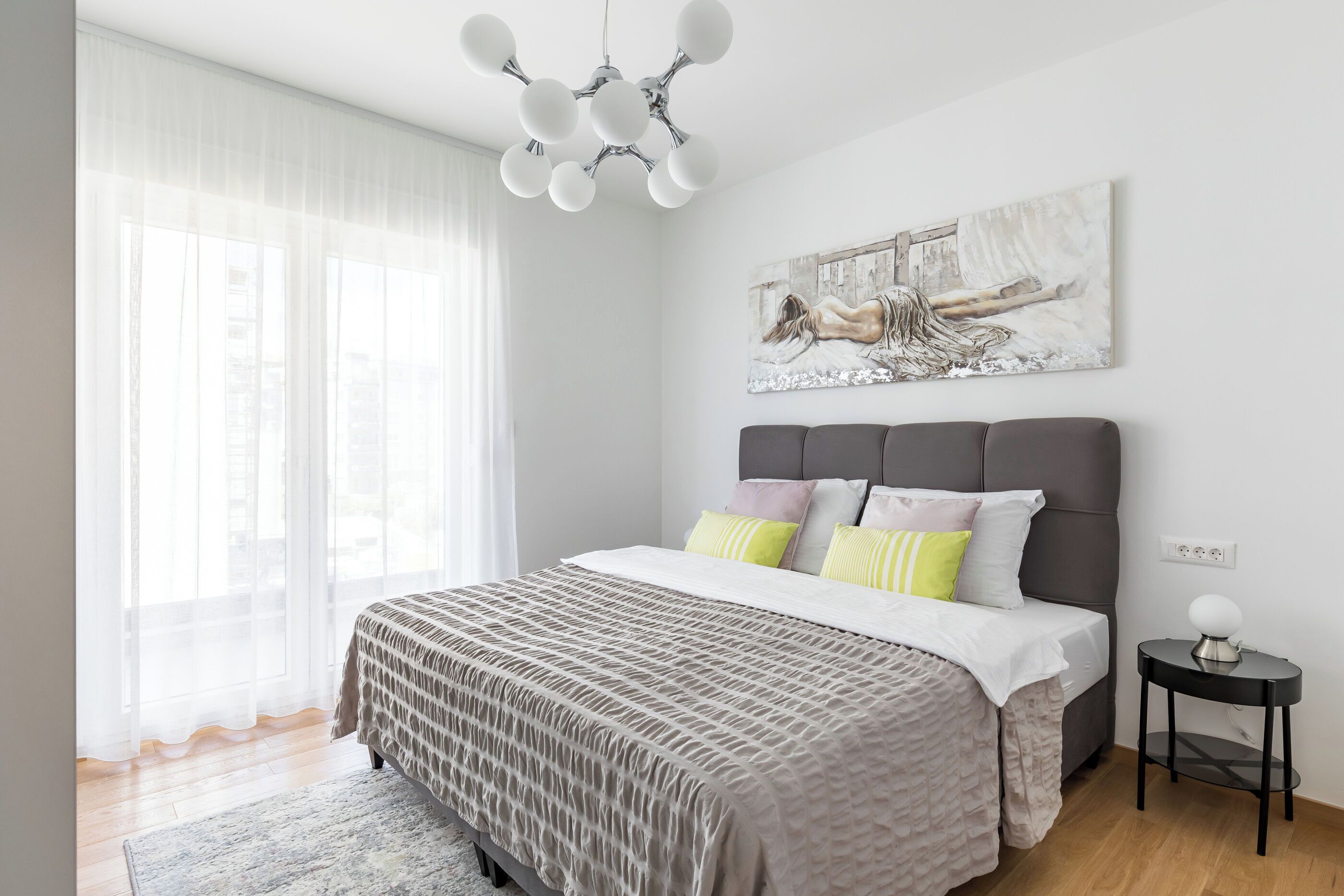 A bedroom with a bed, a bedside table, a window with sheer curtains, and a wall-mounted artwork.