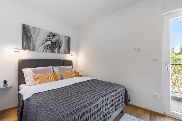 A bedroom with a large bed, a gray headboard, a wall-mounted elephant painting, and a window with a view of buildings. - Apartment Karla (Split)