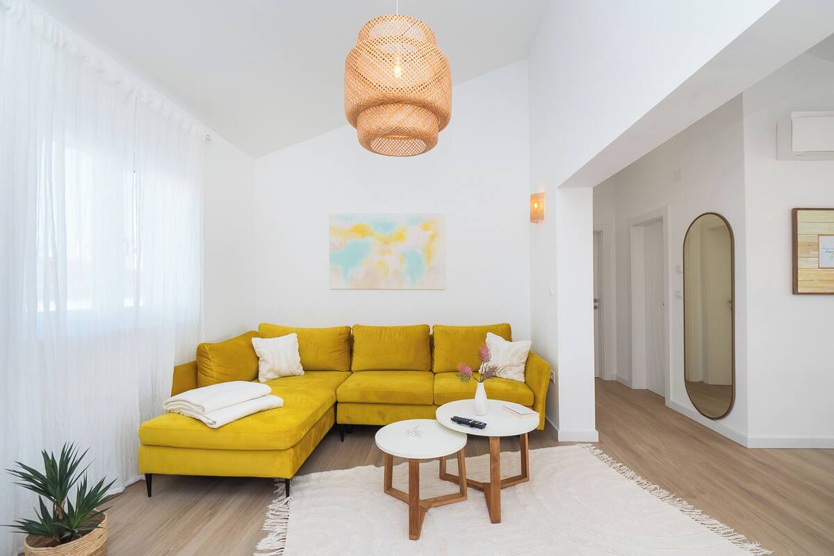 Un salon lumineux avec un canapé d’angle jaune, un tapis blanc et une table ronde en bois.