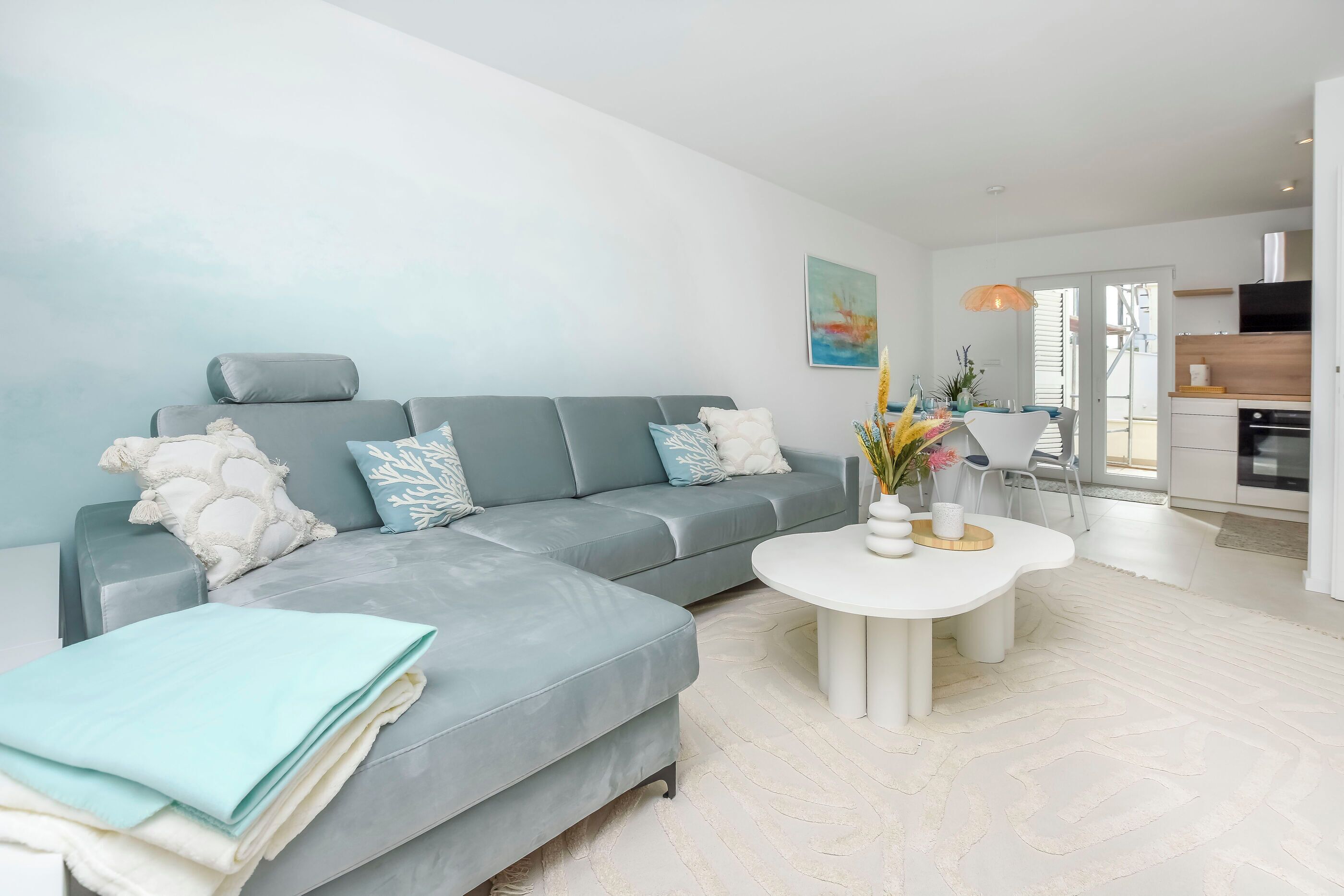A modern living room with a grey sectional sofa, a white coffee table, and a kitchen area visible through an open door.