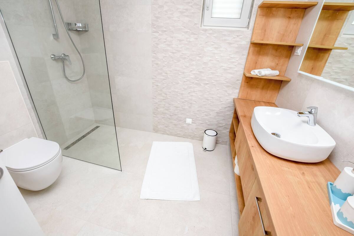 Une salle de bain moderne avec une douche à parois de verre, des toilettes blanches, un meuble-lavabo en bois et une petite étagère avec une serviette.