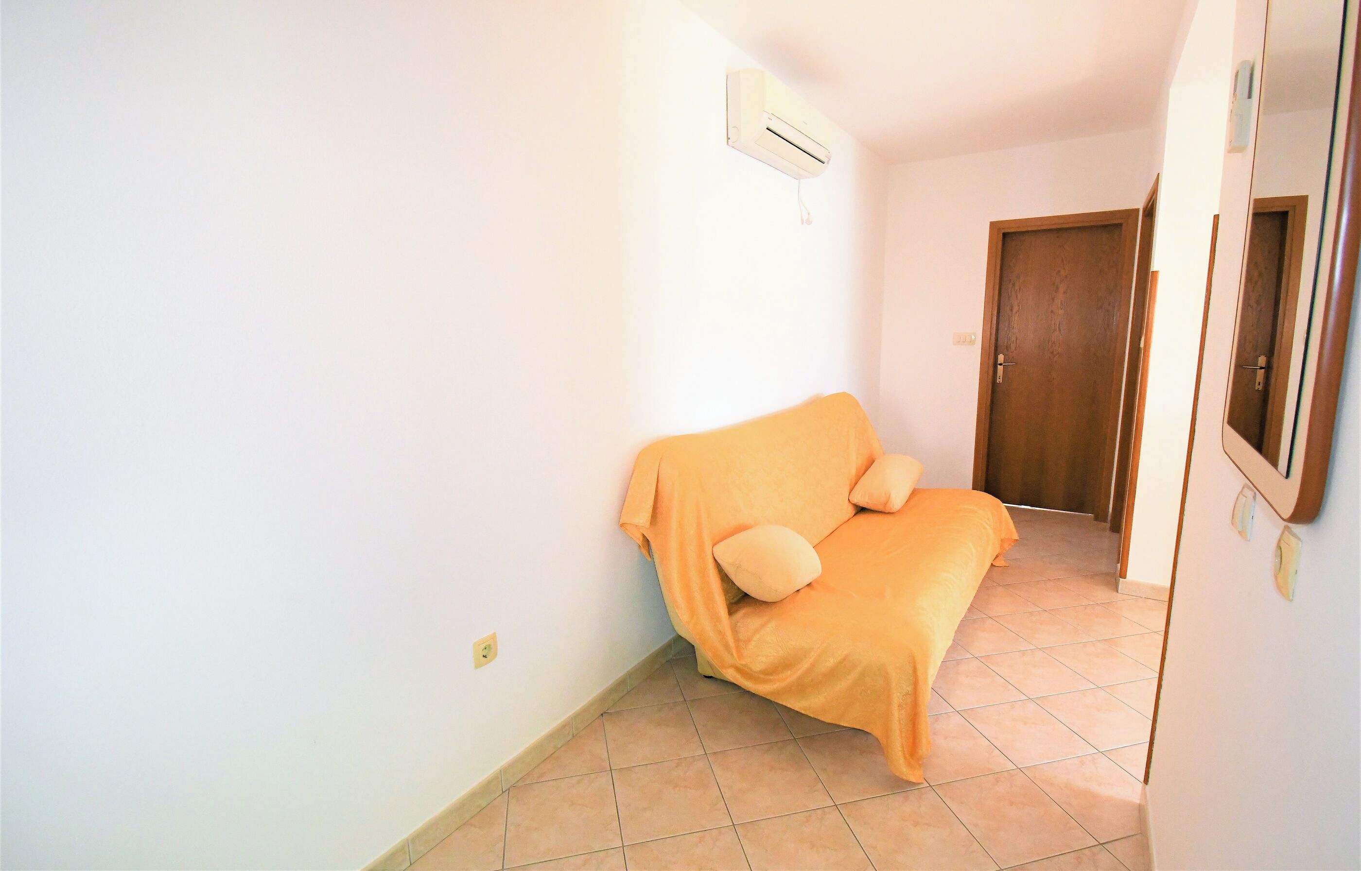A room with a bed covered in an orange blanket, a wooden door, and a white wall with an air conditioning unit.