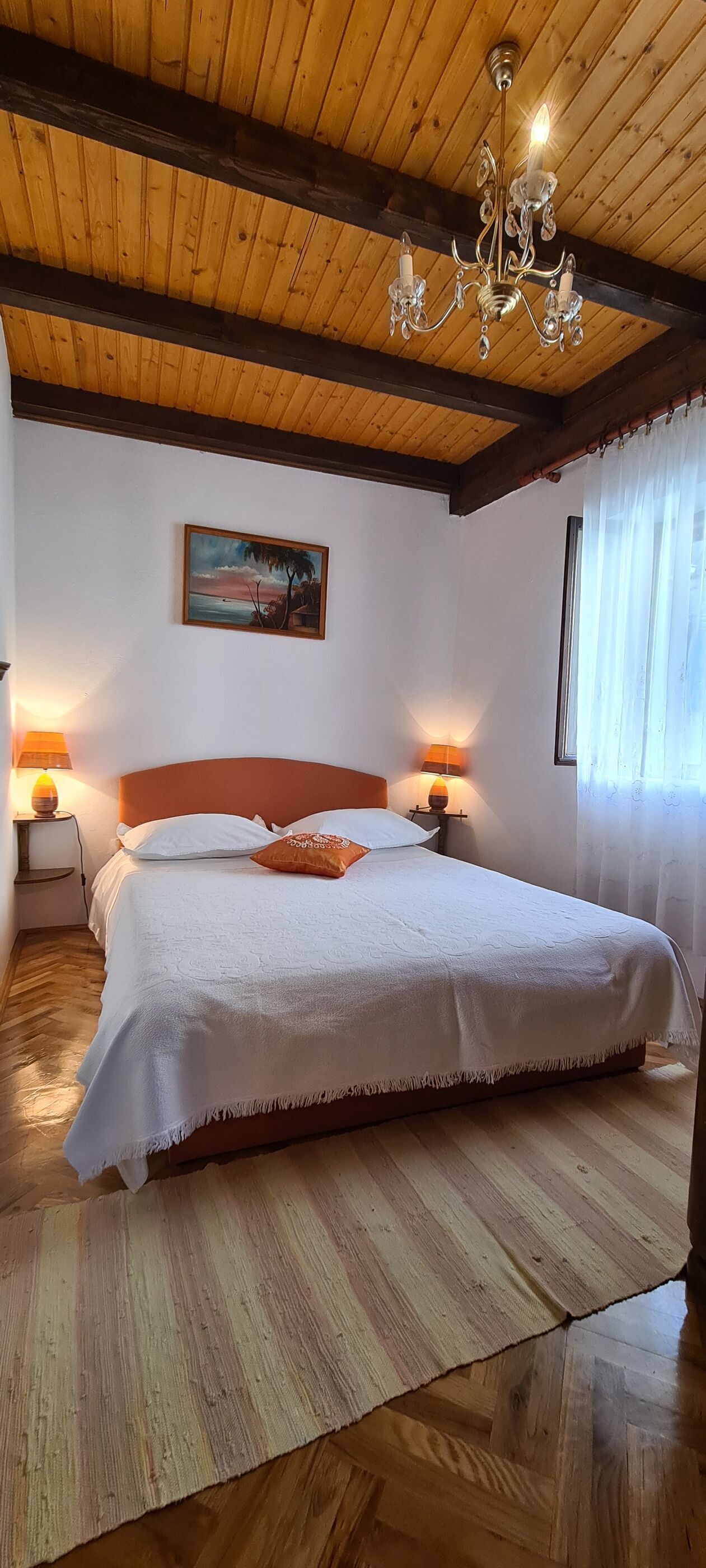 Un dormitori amb sostre de fusta, un lampadari, un llit amb roba de llit blanca, dues làmpades de nit i un quadre a la paret.