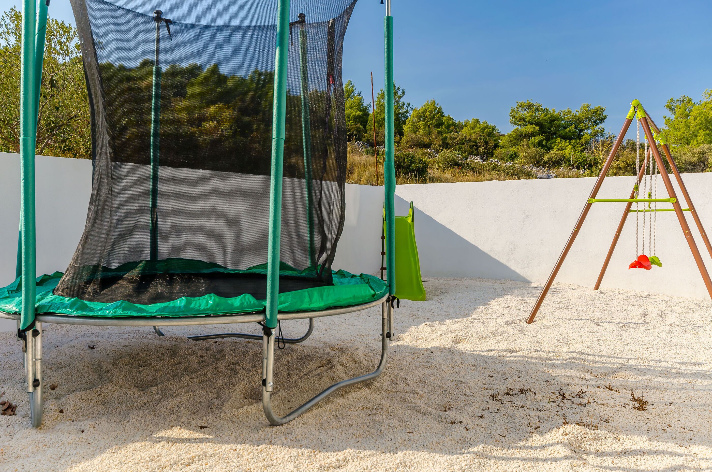 A trampoline with a safety net and a swing set in a sandy area.