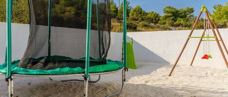A trampoline with a safety net and a swing set in a sandy area.