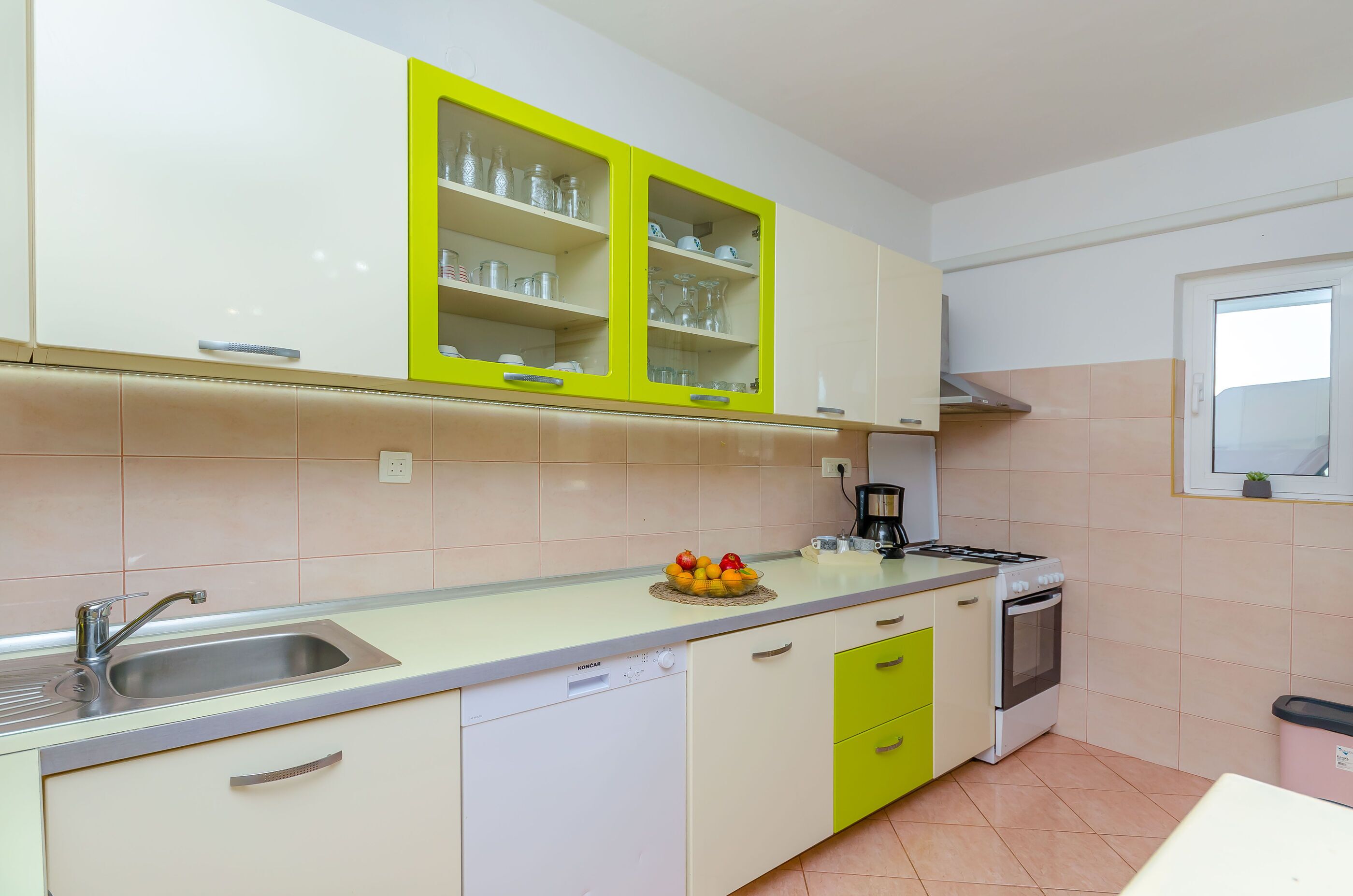 A kitchen with white cabinets, a yellow upper cabinet, a sink, a stove, and a window with a plant.