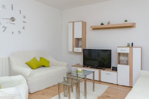 A modern living room with a white sofa, a glass coffee table, a flat-screen TV, and a minimalist shelving unit. - House Roko (Marina)