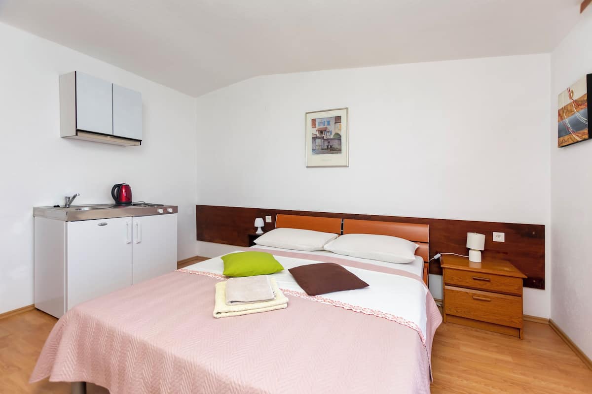 A neatly arranged hotel room with a bed, a small kitchenette, a wooden nightstand, and a framed picture on the wall.