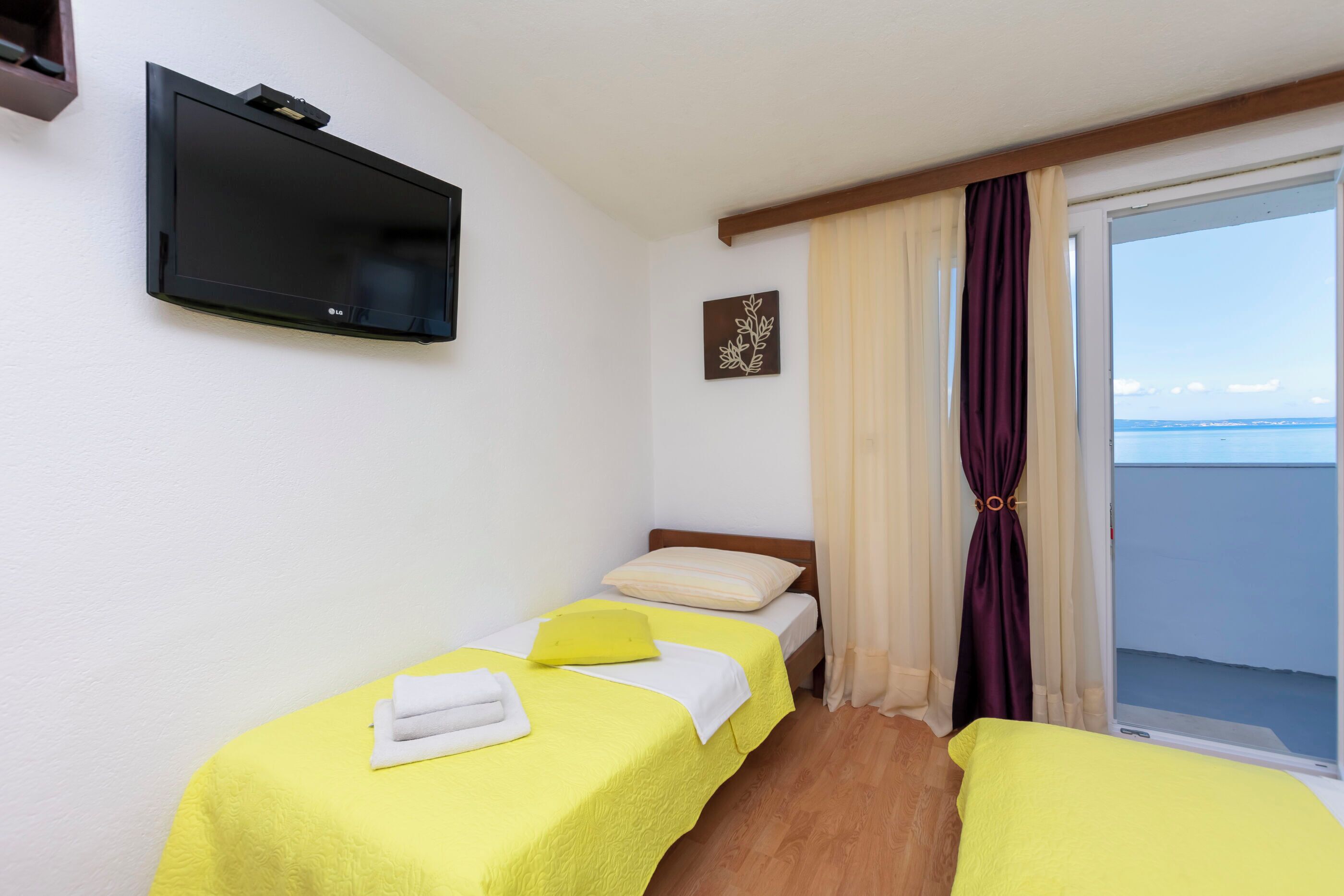 A hotel room with two single beds, a television mounted on the wall, a window with curtains, and a view of the sea.