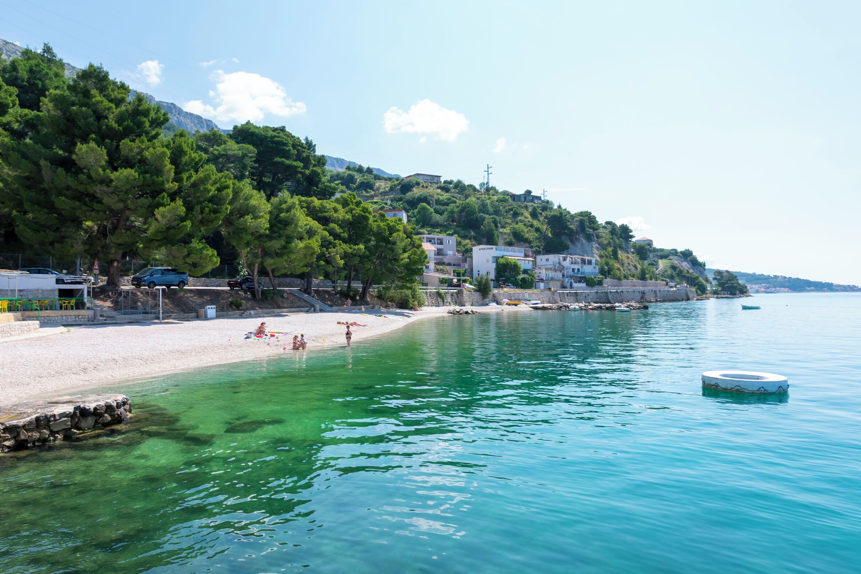 A coastal beach with clear turquoise water, a sandy shore, and buildings on a hillside.