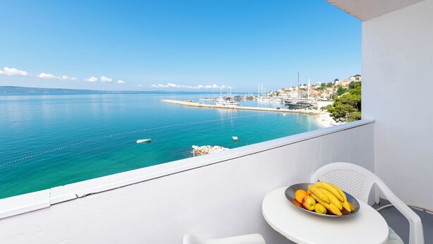 A balcony with a table holding a bowl of fruit, overlooking a coastal town with boats and buildings. - Apartments Luka (Dugi Rat)